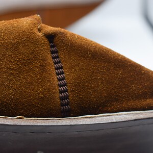 Peut inclure: Gros plan sur une chaussure en daim marron avec une couture marron fonc&eacute; sur le c&ocirc;t&eacute;. La chaussure a un d&eacute;tail de couture blanche autour du bord de la semelle.