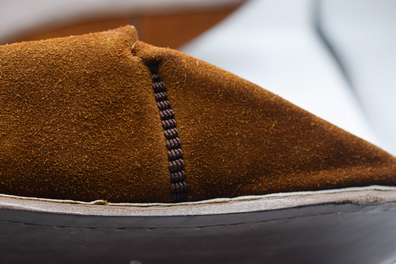 Peut inclure: Gros plan sur une chaussure en daim marron avec une couture marron fonc&eacute; sur le c&ocirc;t&eacute;. La chaussure a un d&eacute;tail de couture blanche autour du bord de la semelle.