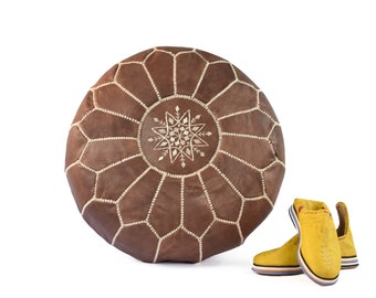 May include: A brown leather pouf with a white star design. The pouf is round and has a tufted surface. Two yellow leather slippers are in the foreground.