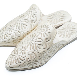 May include: Pair of off-white leather slippers with intricate floral embroidery. The slippers have a pointed toe and a flat sole.