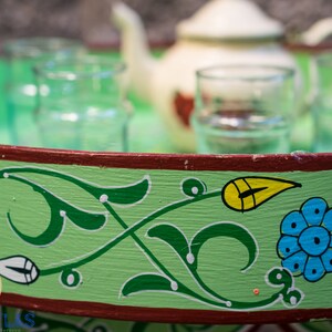 May include: A green wooden tray with a red rim and painted floral designs in green, yellow, blue, and white. The tray has four glasses and a white teapot on it.