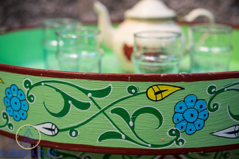 May include: A green wooden tray with a red rim and painted floral designs in green, yellow, blue, and white. The tray has four glasses and a white teapot on it.