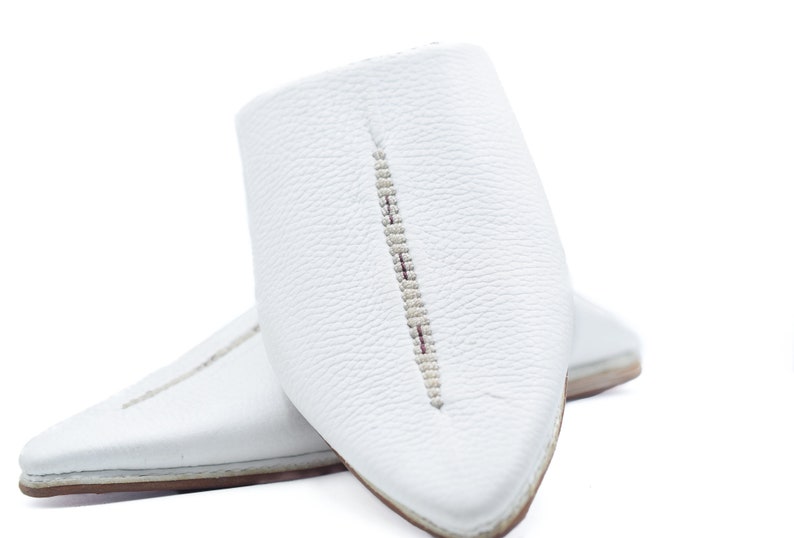 May include: A pair of white leather slippers with pointed toes and a decorative stitch down the center of each shoe.
