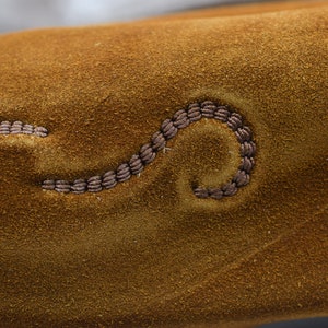 Peut inclure: Gros plan sur une surface en daim marron avec un motif d&eacute;coratif cousu. La couture est en forme de ligne courbe.