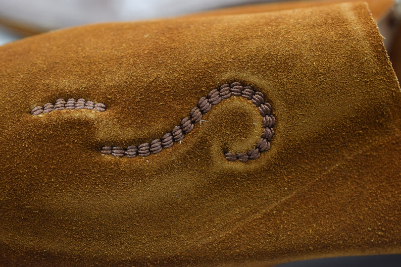Peut inclure: Gros plan sur une surface en daim marron avec un motif d&eacute;coratif cousu. La couture est en forme de ligne courbe.