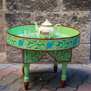 May include: A green painted wooden table with a round top and four legs. The table is decorated with a floral design in green, blue, and yellow. A white teapot and several glass cups are on the table.