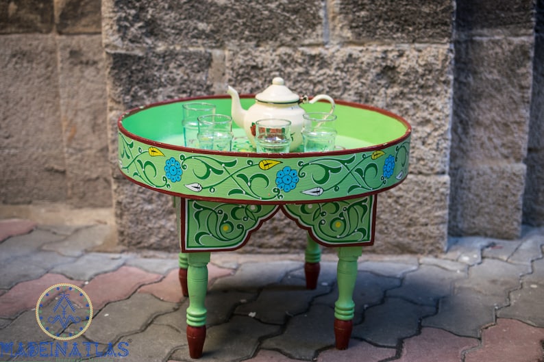 May include: A green painted wooden table with a round top and four legs. The table is decorated with a floral design in green, blue, and yellow. A white teapot and several glass cups are on the table.
