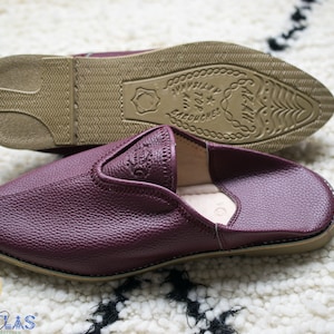 May include: A pair of burgundy leather slippers with a pointed toe and a brown leather sole. The sole has a decorative pattern and the text "MADE IN ATLAS" is embossed on the sole.