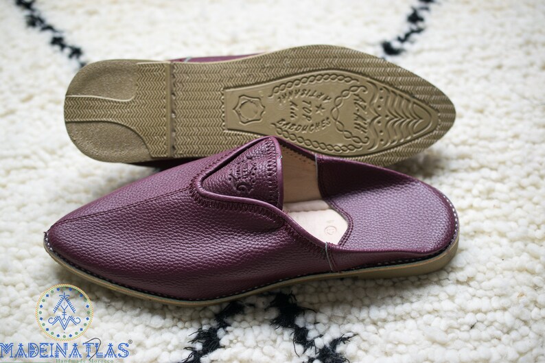 May include: A pair of burgundy leather slippers with a pointed toe and a brown leather sole. The sole has a decorative pattern and the text "MADE IN ATLAS" is embossed on the sole.