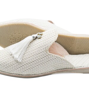 Puede incluir: Un par de zapatillas de color blanco roto con un borla blanca y un adorno de strass. Las zapatillas tienen una parte superior de tejido texturizado y una suela de goma.