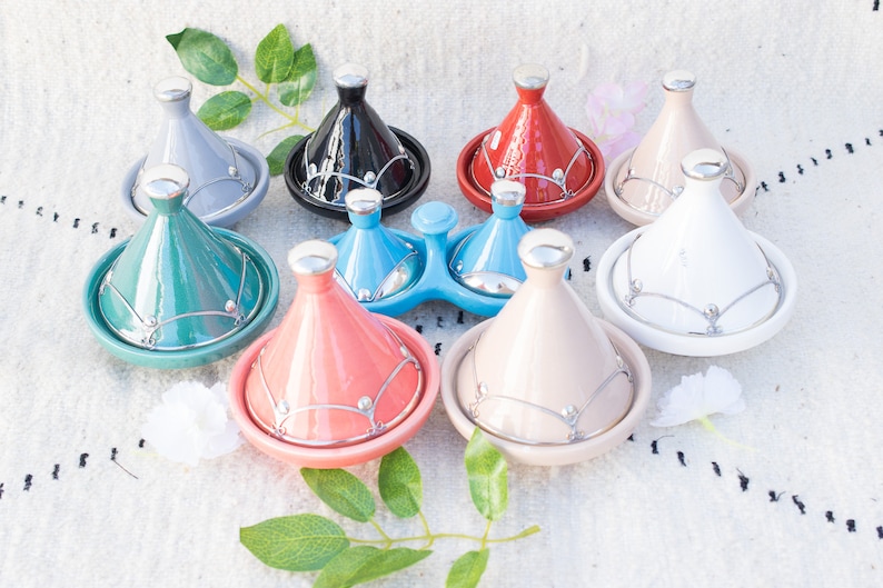 May include: A set of nine colorful ceramic tagines with silver accents. The tagines are arranged in a circle on a white surface.