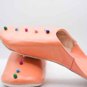 Op de afbeelding: Koraalroze leren slippers met een ronde neus en hielopening. De slippers zijn versierd met kleine, kleurrijke pompons langs de bovenrand. De zool is gebroken wit. Leren slippers voor binnenshuis.