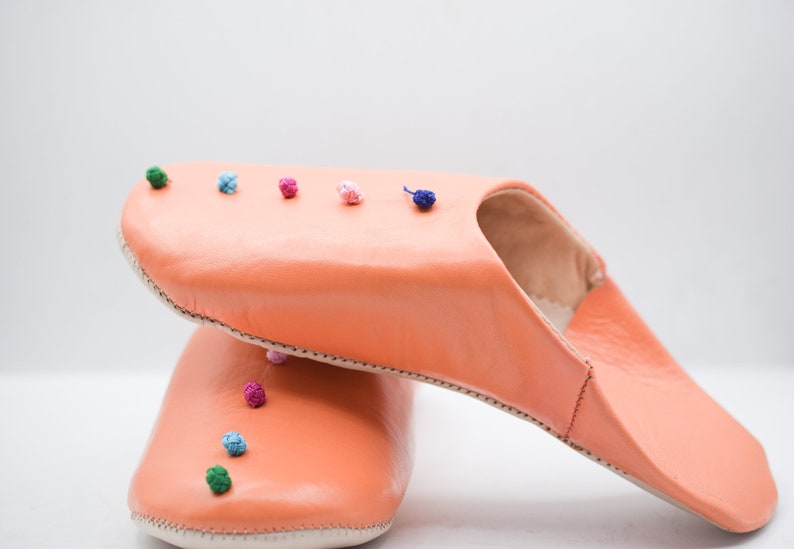 Op de afbeelding: Koraalroze leren slippers met een ronde neus en hielopening. De slippers zijn versierd met kleine, kleurrijke pompons langs de bovenrand. De zool is gebroken wit. Leren slippers voor binnenshuis.