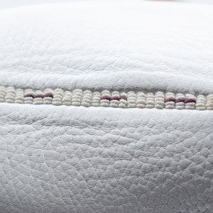 May include: Close-up of a white leather surface with a decorative stitch detail. The stitch is made of white beads with a thin line of purple thread running through the center.