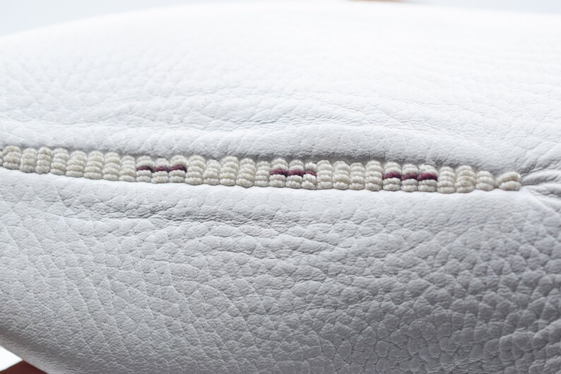 May include: Close-up of a white leather surface with a decorative stitch detail. The stitch is made of white beads with a thin line of purple thread running through the center.