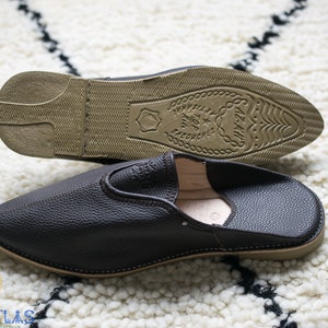 May include: A pair of black leather slippers with a brown leather sole. The sole has the text "Travel Top" and "Morocco" printed on it.