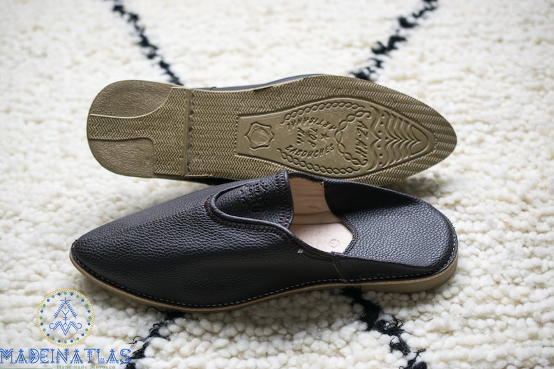 May include: A pair of black leather slippers with a brown leather sole. The sole has the text "Travel Top" and "Morocco" printed on it.
