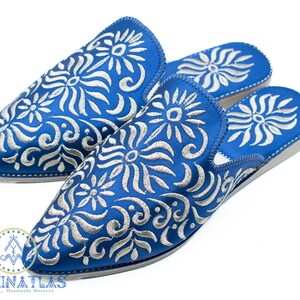 May include: A pair of blue embroidered slippers with a white floral pattern. The slippers are made in Morocco and have a pointed toe.