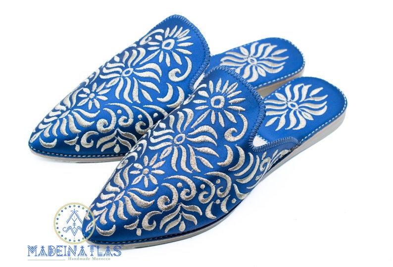 May include: A pair of blue embroidered slippers with a white floral pattern. The slippers are made in Morocco and have a pointed toe.