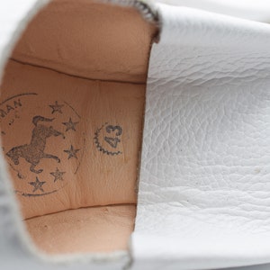 May include: Close-up of the inside of a white leather slipper with a brown leather insole. The insole has a logo with a horse and stars, and the text "43" is printed on the insole.