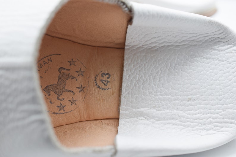 May include: Close-up of the inside of a white leather slipper with a brown leather insole. The insole has a logo with a horse and stars, and the text "43" is printed on the insole.