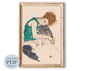 May include: Cross stitch pattern of a woman sitting with her arms around her legs, wearing a green tank top and black trousers. The woman has red hair and is looking down. The background is a light beige colour.