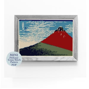 May include: A framed cross stitch pattern of Mount Fuji, a famous Japanese mountain, with a blue sky and white clouds. The mountain is red, green, and blue. The frame is gray.