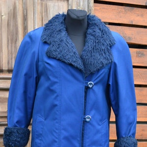 May include: A long, royal blue coat with a faux fur collar and cuffs. The coat has a button-up front and is displayed on a black mannequin. The background features a wooden door and fence.