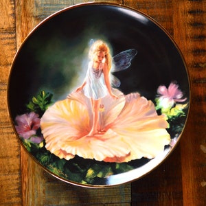 May include: A round decorative plate with a black background featuring a central illustration of a small figure with translucent wings standing on a large, pale yellow flower. The plate has a thin gold-colored rim and includes green foliage.