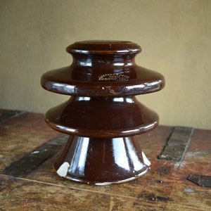 May include: A dark brown glazed ceramic electrical insulator with a tiered, bell-shaped profile. The object features a smooth, reflective surface and embossed text on the upper section that reads 1920 and other characters, with minor chips at the base.