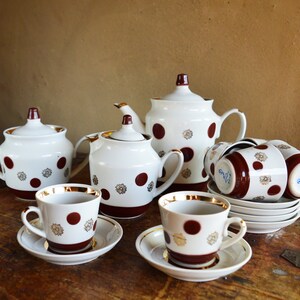 May include: A white porcelain tea set featuring a pattern of dark red polka dots and gold floral accents. The set includes a teapot, a lidded sugar bowl, a creamer, and several teacups with matching saucers, all finished with gold rims.