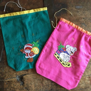 Könnte beinhalten: Zwei Stoffbeutel mit Kordelzug, einer in Grün mit gelbem Rand oben und einem gestickten Weihnachtsmann mit Baum, der andere in Pink mit orangefarbenem Rand oben und einem gestickten weißen Bären auf einem gelben Schlitten, mit goldenen Sternen.