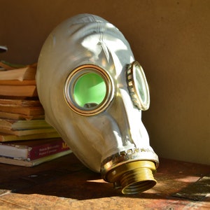 Op de afbeelding: Een wit gasmasker met groene ooglenzen en een messing mondstuk. Het masker ligt op een houten oppervlak.