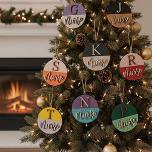 Peut inclure: Un sapin de Noël décoré de plusieurs ornements ronds. Chaque ornement est divisé en deux sections avec une lettre et le mot "Nurse". Les ornements sont de différentes couleurs, dont le violet, le noir et le jaune.