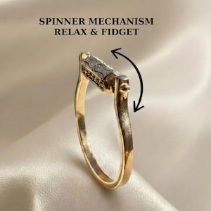 May include: A gold-toned ring featuring a central rectangular spinner mechanism with a textured surface. The text SPINNER MECHANISM RELAX & FIDGET is printed above the ring, accompanied by a curved black arrow indicating the rotational movement.