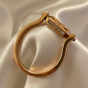 Könnte beinhalten: Ein roségoldener Ring mit einem rechteckigen Mittelstück, das mit kleinen, funkelnden Diamanten besetzt ist. Der Ring hat ein einzigartiges Design mit einem gebogenen Band und einer geometrischen Form, vor weißem Satin.