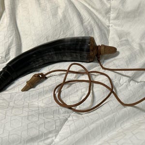 Puede incluir: Un recipiente en forma de cuerno negro y gris con un tapón de madera y un cordón de cuero marrón. El recipiente tiene un acabado oscuro y pulido y una pieza decorativa de madera unida al cordón.