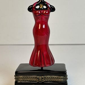 Puede incluir: Adorno de vestido rojo sobre una caja negra con detalles dorados. El vestido es de un rojo vibrante con una parte superior negra y un acento dorado. La caja lleva la firma "Marilyn Monroe" en oro.