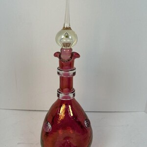 May include: A ruby red glass bottle with a clear glass stopper. The bottle has a bulbous shape with textured detailing and two decorative circular accents. The stopper is elongated with a rounded top and a clear glass tip.