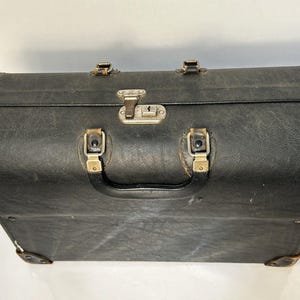 Pode incluir: Mala preta vintage com ferragens metálicas, incluindo uma fechadura central, protetores de canto e alça. A mala tem uma superfície texturizada e parece ser feita de couro ou material semelhante a couro. As ferragens têm um acabamento dourado.