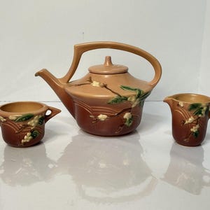 May include: A vintage tea set in shades of peach and brown, featuring a teapot with a curved handle, a sugar bowl, and a creamer. The set is decorated with floral accents in white and green.