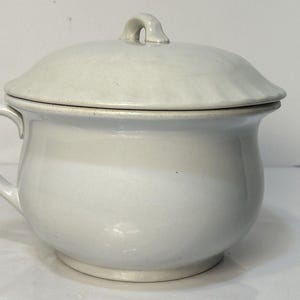 May include: A white ceramic chamber pot with a matching lid. The pot has a rounded body, a handle, and a lid with a small loop. The piece is likely vintage and has a simple, functional design.