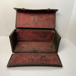 May include: An open, antique, brown leather box with a red velvet interior. The box has a hinged lid and a rectangular shape. The interior shows signs of wear and age. The box is empty and sits on a white surface.
