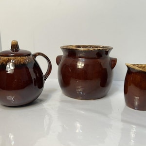 May include: A set of three brown ceramic kitchen items: a teapot with a lid, a small pot with handles, and a creamer. The items have a glossy finish with a lighter brown trim around the edges.