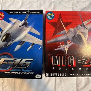 May include: Two video game boxes featuring fighter jets. The box on the left is blue with an F-16 fighter jet and the text "F-16 MULTIROLE FIGHTER." The box on the right is red with a MiG-29 fighter jet and the text "MiG-29 FULCRUM."