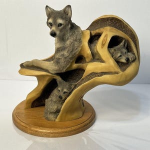 May include: A detailed sculpture of a wolf family. The sculpture features a mother wolf and two cubs nestled within a stylized, organic form. The base is a round, wooden platform. The wolves are depicted in shades of gray and brown.