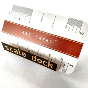 May include: A white rectangular card with rounded corners featuring a brown horizontal stripe with the text ARC CARRY in white. The card includes printed imperial ruler markings in inches along the top and bottom edges, with a dark brown rectangular piece attached to the front displaying the text scale dock in a light-colored sans-serif font.