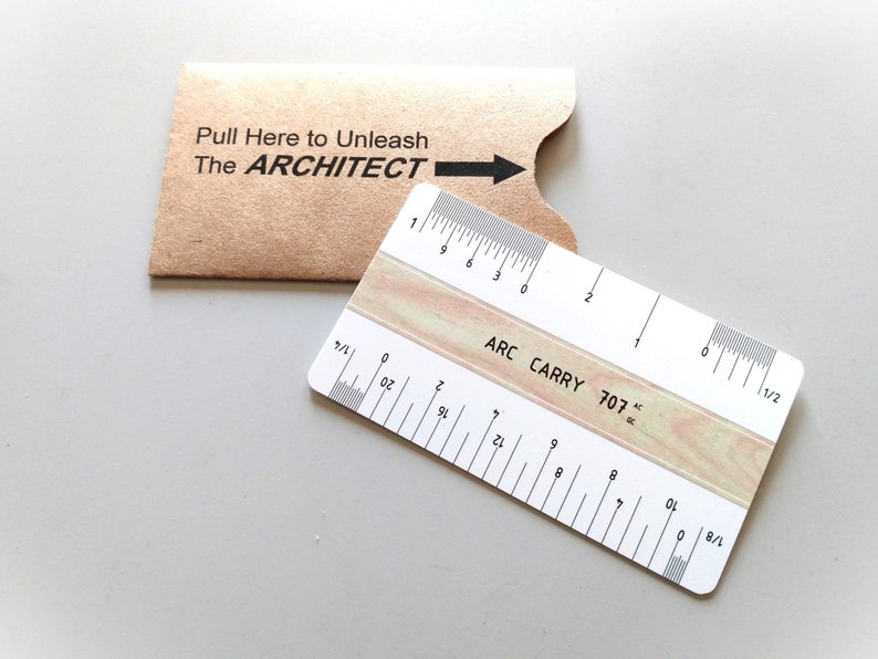 May include: A white ruler with black markings labeled 'ARC CARRY 707' and '1/2', '1/4', '1/8', and '1/16'. The ruler is on a light gray surface and is partially covered by a brown envelope with the text 'Pull Here to Unleash The ARCHITECT' printed on it.