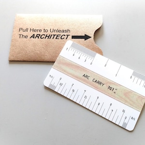 May include: A white and beige ruler with black markings and the text "ARC CARRY 707". The ruler is next to a brown card with the text "Pull Here to Unleash The ARCHITECT" and an arrow.