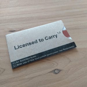 May include: A rectangular card with the text "Licensed to Carry" in black. The card is light brown with a black stripe at the bottom containing text. The top right corner has a red accent. The card is on a wooden surface.